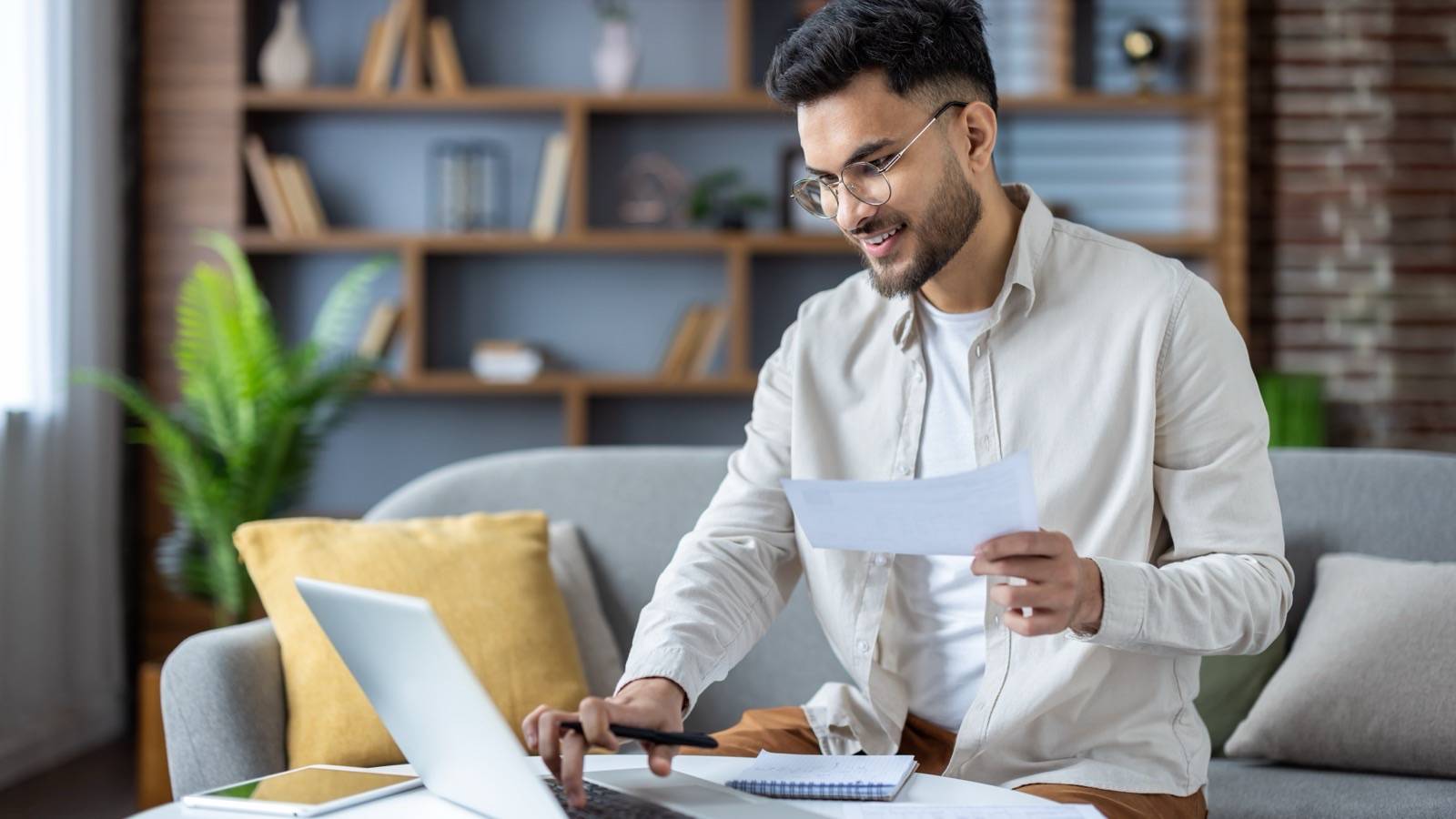 Man typing on a laptop at home, holding a document