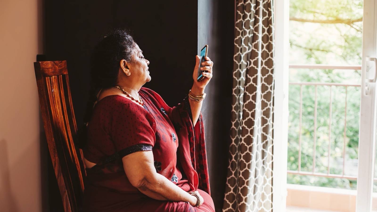 Older woman in a sari holding up her phone by a window at home