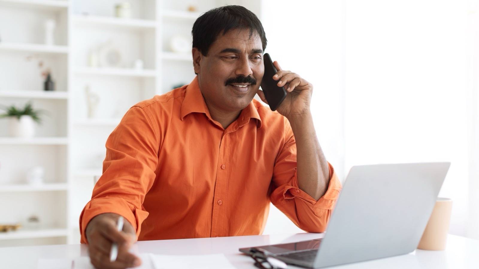 Man on a phone call at home, taking notes beside a laptop